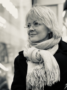 Vaaleatukkainen nainen katsoo mietteliäänä sivulle yläviistoon takki päällä ja kaulahuivi kaulassa.