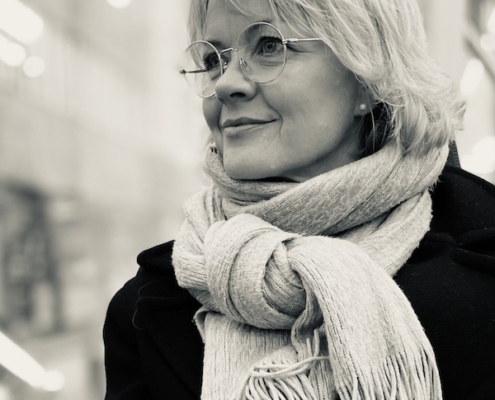Vaaleatukkainen nainen katsoo mietteliäänä sivulle yläviistoon takki päällä ja kaulahuivi kaulassa.