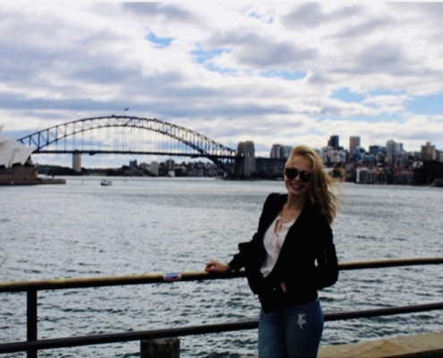 Nuori nainen Sydneyn oopperatalon edustalla hiukset tuulessa hulmuten.