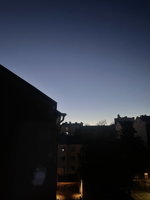 Aamunäkymä ikkunasta, kun on vielä pimeää ja näkyy vain talojen mustat ääriviivat aamutaivasta vasten.