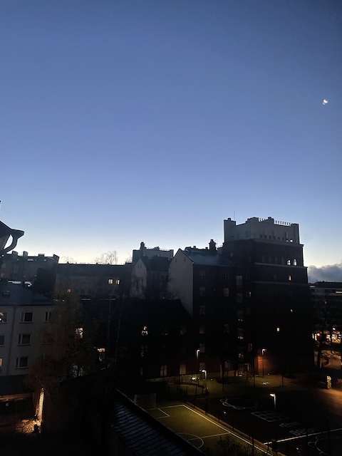 Aamunäkymä ikkunasta, kun on vielä pimeää ja näkyy vain talojen mustat ääriviivat aamutaivasta vasten.